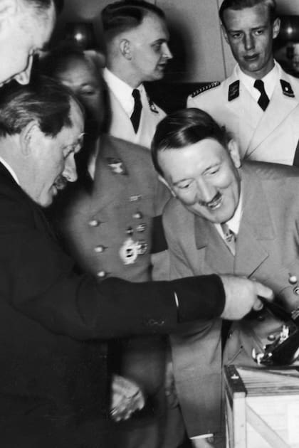 Adolf Hitler observa una maqueta del "auto del pueblo" y se ríe al descubrir al motor en la parte trasera. Quien levanta la tapa del baúl es el diseñador Ferdinand Porsche