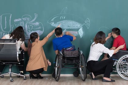 Group of special students in classroom, a down syndrome girl, two handicapped boys and  two female teachers drawing and painting on black board together.