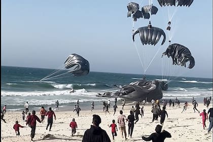 Palestinos corriendo hacia paracaídas sujetos a paquetes de alimentos, lanzados desde aviones estadounidenses en una playa de la Franja de Gaza el 2 de marzo de 2024