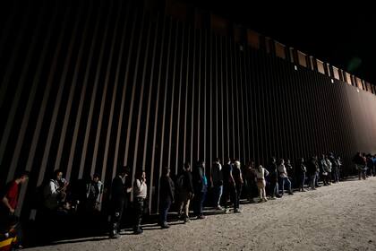 Un grupo de personas forman una fila junto a un muro fronterizo mientras esperan para solicitar asilo tras cruzar la frontera desde México, el 11 de julio de 2023, cerca de Yuma, Arizona (Archivo).