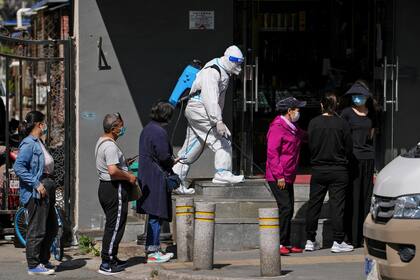 Un operario equipado con un traje de protección rocía desinfectante mientras residentes, con mascarilla, esperan en fila para someterse a una prueba de detección del coronavirus en un complejo residencial, el 3 de mayo de 2022, en Beijing. (AP Foto/Andy Wong)